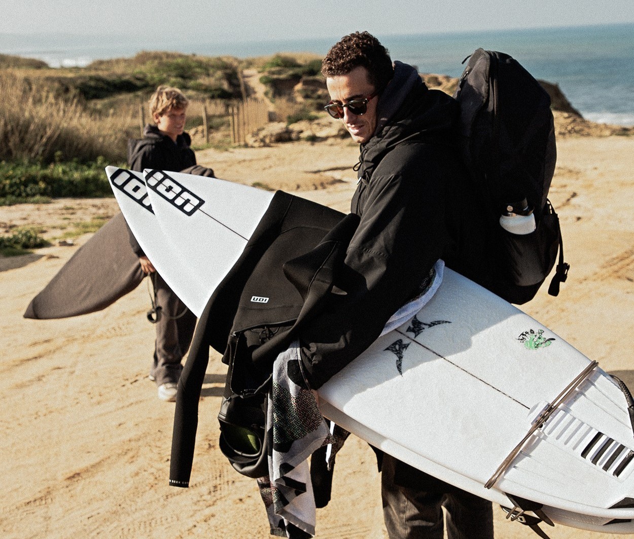 Surfeur transportant sa combinaison néoprène ION et sa planche sur la plage