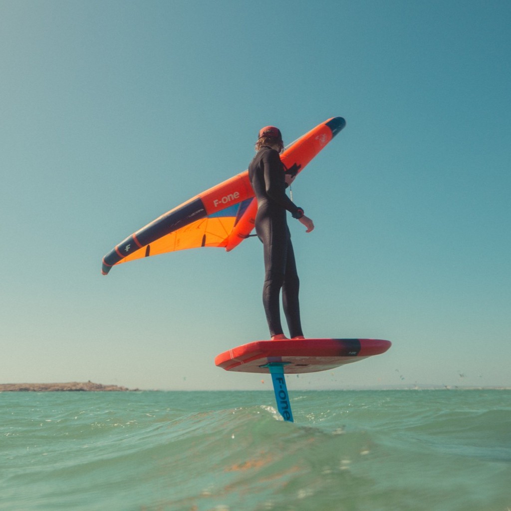 Rideuse en pleine session de wingfoil avec l'aile F-One Origin sur une mer calme, avec la côte en arrière-plan.