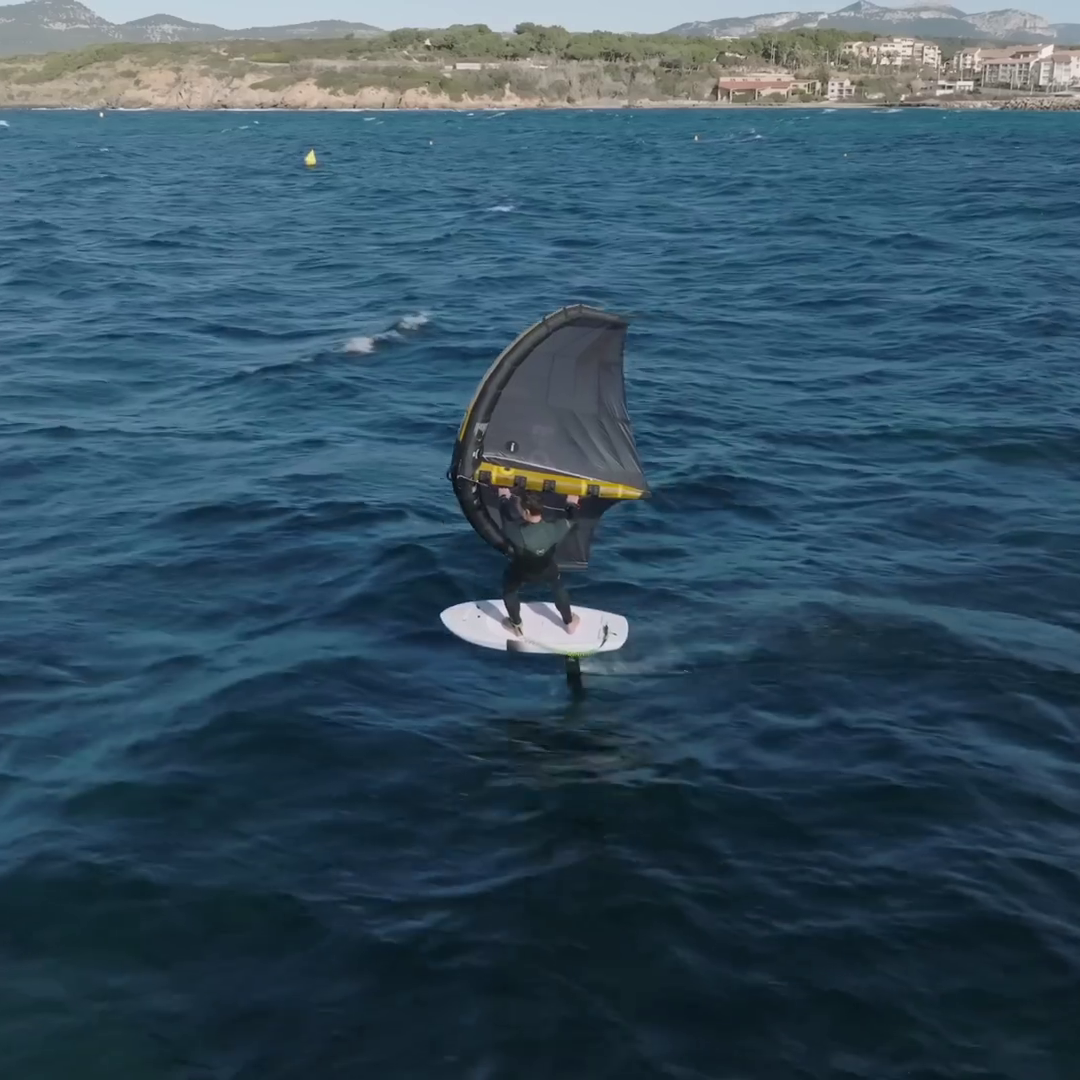 Rider pratiquant le wingfoil sur une mer agitée avec une aile Strike V5 noire et jaune et une planche foil, avec un littoral en arrière-plan.