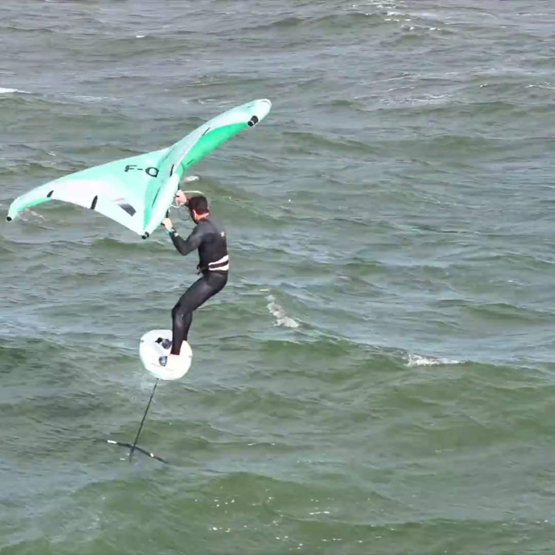 Rider en combinaison noire effectuant un saut en wingfoil au-dessus d'une mer agitée, avec une aile F-One Strike V5 verte et blanche.