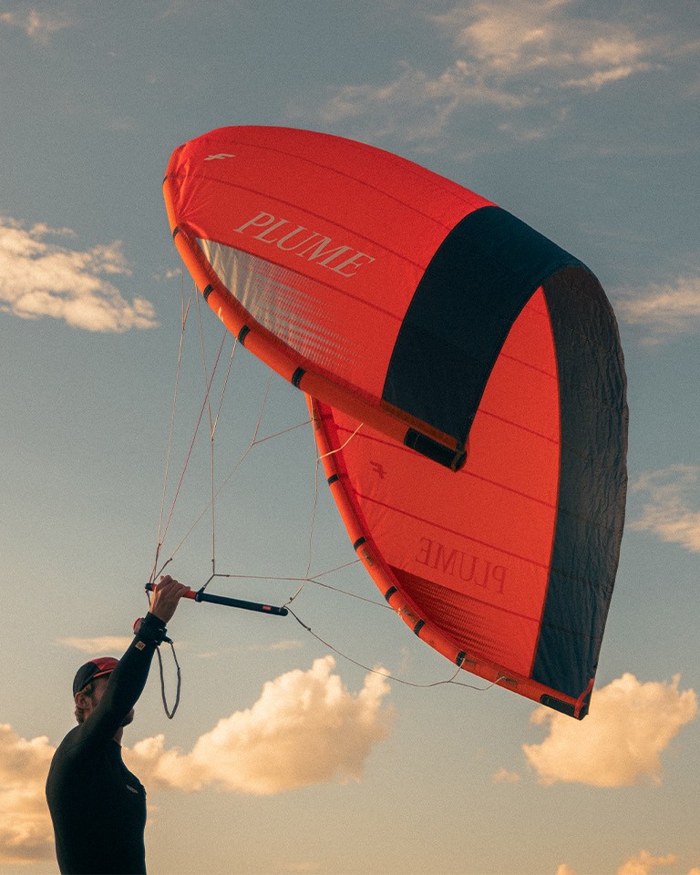 Personne tenant une aile de parawing F-One Plume rouge et noire, ciel dégagé avec quelques nuages en arrière-plan.