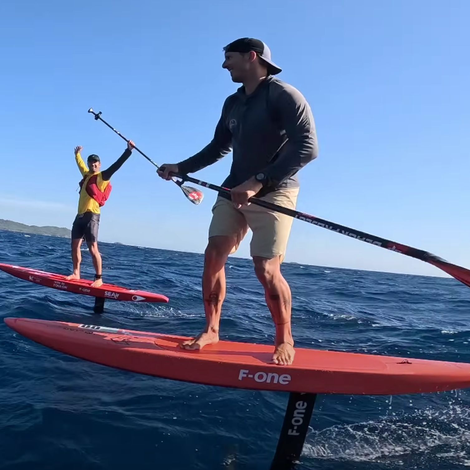 Deux riders en SUP foil F-One naviguant en downwind, pagaie à la main, sur mer bleue avec du vent